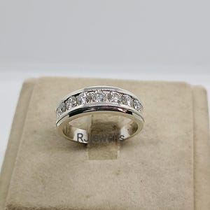 May include: A silver ring with a row of clear gemstones set across the top. The ring has a polished finish and a simple, elegant design. The ring is displayed on a neutral-toned surface.