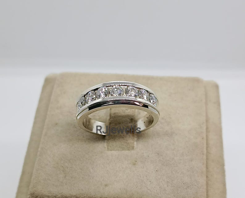 May include: A silver ring with a row of clear gemstones set across the top. The ring has a polished finish and a simple, elegant design. The ring is displayed on a neutral-toned surface.
