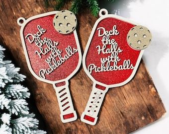 May include: Two red and white wooden pickleball paddle ornaments with glitter. The paddles have the text "Deck the Halls with Pickleballs" and one paddle has the text "Joe & Margie".