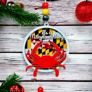 Peut inclure: Une décoration de Noël blanche et argentée avec une silhouette de crabe rouge et le texte "It's a Maryland thing" sur un fond blanc avec les couleurs du drapeau de l'État du Maryland.