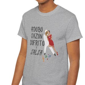 Puede incluir: Una camiseta gris con las palabras "Adobo, Sazon, Sofrito y Salsa" en escritura negra. La camiseta también tiene un gráfico de una pareja bailando en blanco y rojo, con notas musicales.