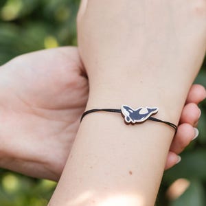 Könnte beinhalten: Ein schwarzes Armband mit einem Schmetterlingsanhänger. Der Schmetterlingsanhänger ist schwarz und weiß. Das Armband wird an einem Handgelenk vor grünem Laub getragen.