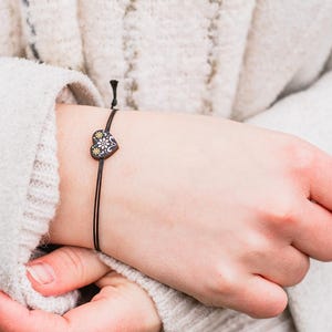 Könnte beinhalten: Nahaufnahme eines Handgelenks, das ein schwarzes Armband aus Schnur mit einem kleinen silbernen Herz-Anhänger trägt.
