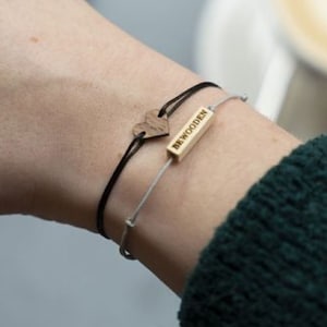 Könnte beinhalten: Eine Person trägt einen grünen Pullover mit zwei Armbändern am Handgelenk. Ein Armband ist schwarz mit einem kleinen Herz-Anhänger und das andere ist silber mit einem Balken-Anhänger, auf dem "remarkable" steht.