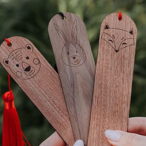 Könnte beinhalten: Drei Holz-Lesezeichen mit Tierillustrationen. Die Lesezeichen sind braun mit einem roten Quasten an einem. Die Lesezeichen zeigen einen Bären, ein Kaninchen und einen Fuchs. Der Text "Lissarna" ist auf das Fuchs-Lesezeichen graviert.