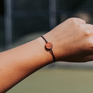 Könnte beinhalten: Ein schwarzes Armband aus Schnur mit einem kleinen, basketballförmigen Holzanhänger.