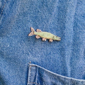Könnte beinhalten: Eine hellgrüne und rosa Fisch-Emaille-Anstecknadel ist an einer blauen Jeansjacke befestigt. Die Anstecknadel ist mit Flossen und Schuppen versehen. Die Jacke hat eine Knopfleiste und eine Brusttasche. Der Hintergrund ist unscharf.
