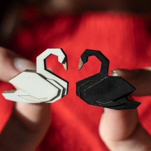 Può includere: Due cigni di origami, uno bianco e uno nero, sono tenuti dalle dita di una persona. I cigni si guardano, creando una forma di cuore.