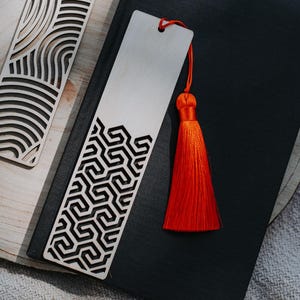 Könnte beinhalten: Zwei Holz-Lesezeichen mit aufwendigen Ausschnitten und Quasten. Eines hat eine schwarze Quaste und ein wellenförmiges Muster, das andere eine orangefarbene Quaste und ein geometrisches Design. Sie liegen auf einer Holzoberfläche neben einem geschlossenen schwarzen Buch.