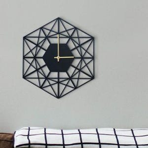 Op de afbeelding: Een zwarte geometrische klok met gouden wijzers hangt aan een witte muur boven een bruine bank met zwart-witte gepatroneerde kussens en een zwart-wit geblokte deken.
