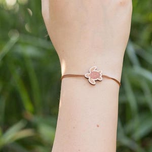 Può includere: Un braccialetto con cordino marrone con un piccolo ciondolo a forma di tartaruga marrone chiaro. Il braccialetto è indossato su un braccio, con uno sfondo verde sfocato di fogliame.