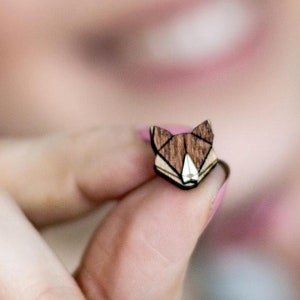 Könnte beinhalten: Eine kleine Holz-Fuchs-Kopf-Anstecknadel aus braunem und hellbraunem Holz wird in der Hand einer Person gehalten. Die Anstecknadel ist geometrisch gestaltet.