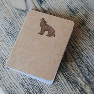 May include: A brown notebook with a wooden wolf cutout on the front cover. The notebook is sitting on a wooden table.