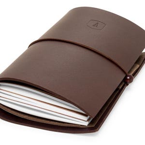 May include: A brown leather bound journal with a leather strap closure and a small wooden bead. The journal has a small logo on the front cover.