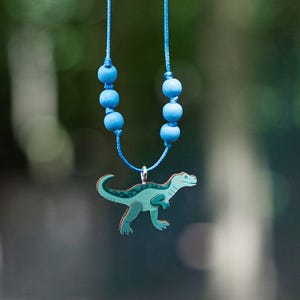Könnte beinhalten: Eine blaue Halskette mit einem Dinosaurier-Anhänger. Die Halskette hat blaue Perlen und eine blaue Schnur.