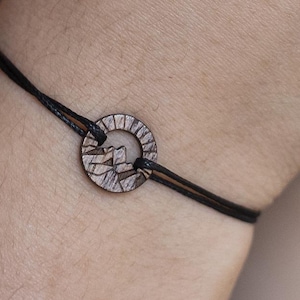 Puede incluir: Un brazalete de cordón negro con un dije de madera con un diseño de montaña tallado.