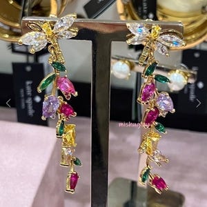 Puede incluir: Pendientes colgantes dorados con diseño floral. Cada pendiente presenta una parte superior en forma de libélula con cristales transparentes y amarillos. Debajo, gemas de colores rosa, morado, verde y amarillo caen en cascada, creando un accesorio vibrante.