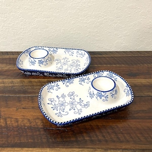 May include: Two rectangular ceramic serving trays with rounded corners, each featuring a white base with blue floral patterns and a built-in circular egg cup. The edges are decorated with a blue and white checkered pattern along the rim.