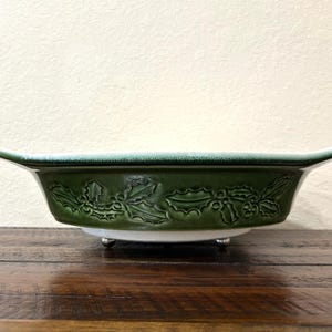 May include: A green ceramic serving dish with a holly leaf and berry design. The dish has a white base and sits on three small silver feet. The rim is slightly flared and has a darker green color.