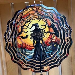 May include: A metal wind spinner with a witch silhouette in front of a full moon and trees. The spinner has a layered design with a black, orange, and yellow color scheme. A beaded heart charm hangs from the bottom.