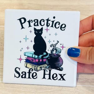 Puede incluir: Posavasos cuadrado blanco con un gato negro sentado sobre libros, un caldero y las palabras "Practice Safe Hex". El diseño incluye estrellas y un tema mágico.