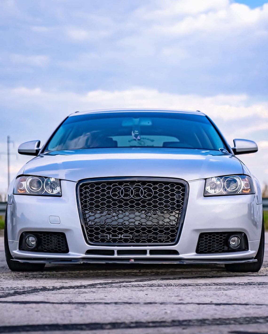 Könnte beinhalten: Ein silberner Audi A3 mit einem schwarzen Wabengrill und einer schwarzen Frontstoßstange. Das Auto steht auf einer grauen Asphaltfläche.