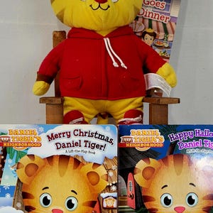 Peut inclure: Une peluche Daniel le Tigre, v&ecirc;tue d'un sweat &agrave; capuche rouge et d'un pantalon jaune, est assise sur une petite chaise en bois. Des livres avec Daniel le Tigre sont devant le jouet. Un livre dit "Joyeux No&euml;l, Daniel le Tigre!"
