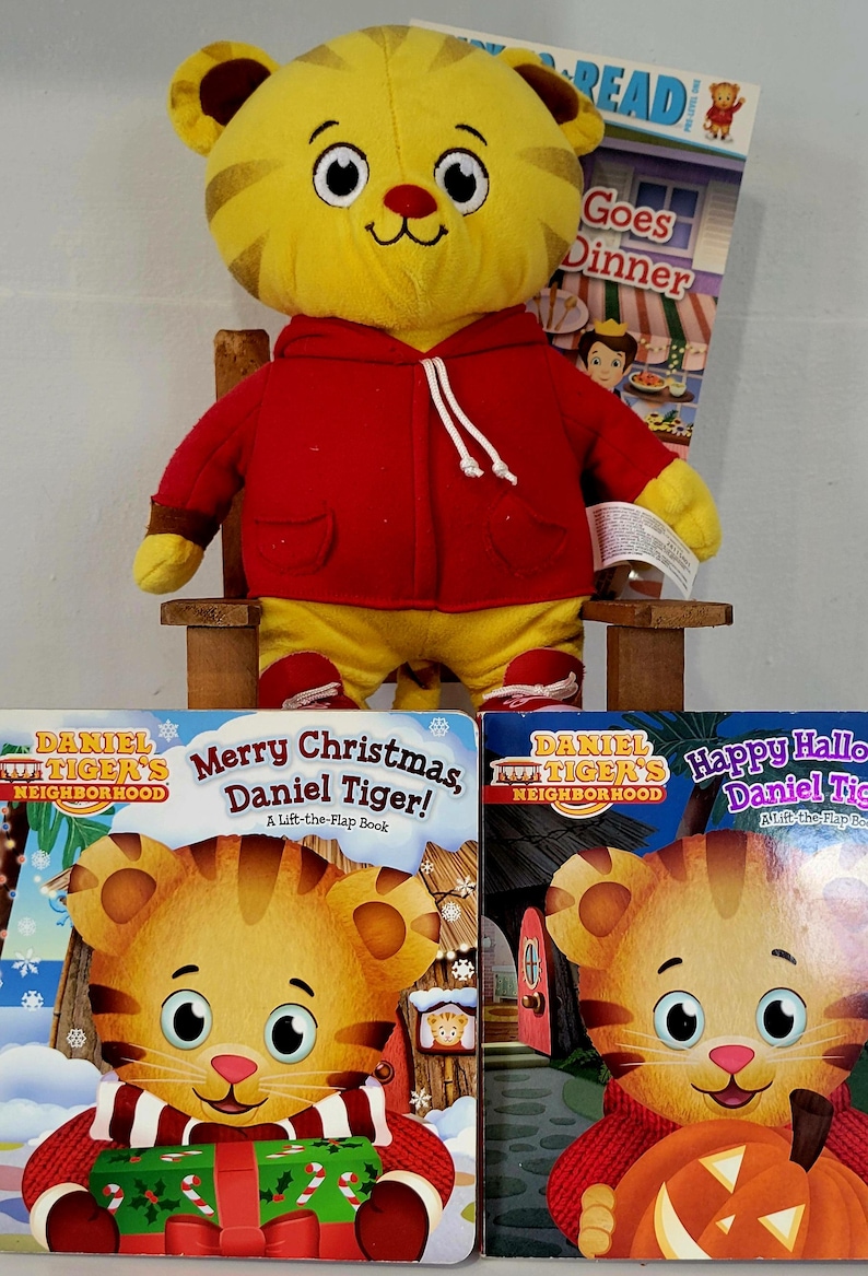 Peut inclure: Une peluche Daniel le Tigre, v&ecirc;tue d'un sweat &agrave; capuche rouge et d'un pantalon jaune, est assise sur une petite chaise en bois. Des livres avec Daniel le Tigre sont devant le jouet. Un livre dit "Joyeux No&euml;l, Daniel le Tigre!"