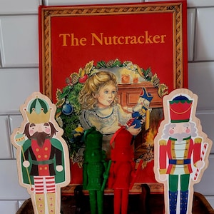 May include: A set of five wooden nutcracker figurines. The figurines are all different colors and designs. The largest figurine is a king with a gold crown and a green cape. The other figurines are a soldier, a green soldier, a red soldier, and a candy cane soldier. The figurines are standing in front of a book titled "The Nutcracker".