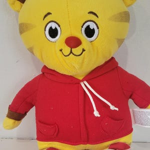 Peut inclure: Peluche repr&eacute;sentant un tigre jaune avec des rayures marron, v&ecirc;tu d'un sweat &agrave; capuche rouge avec des cordons blancs. Le tigre a un nez rouge et des yeux blancs et noirs. Il porte des chaussures blanches et rouges.