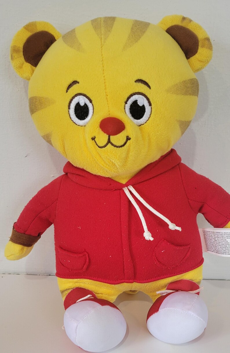 Peut inclure: Peluche repr&eacute;sentant un tigre jaune avec des rayures marron, v&ecirc;tu d'un sweat &agrave; capuche rouge avec des cordons blancs. Le tigre a un nez rouge et des yeux blancs et noirs. Il porte des chaussures blanches et rouges.