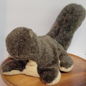 May include: A plush squirrel toy with gray fur and a bushy tail. The toy has a light brown belly and paws, and a small, brown ear. The squirrel is lying on a wooden plate.