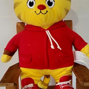 Peut inclure: Peluche repr&eacute;sentant un tigre jaune avec des rayures marron, v&ecirc;tu d'un sweat &agrave; capuche rouge et de baskets rouges. Le tigre a un nez rouge et un visage souriant, assis sur une petite chaise en bois.