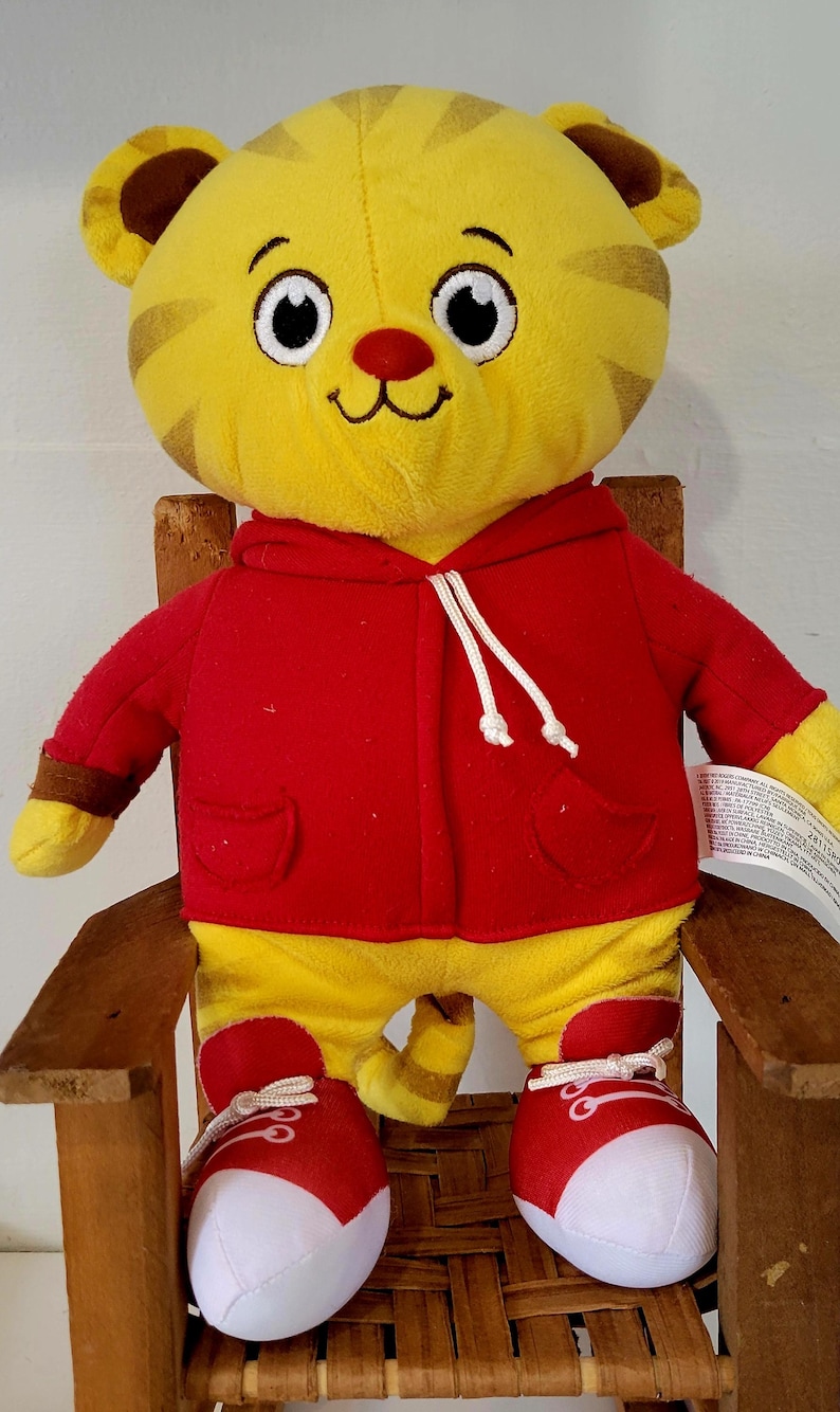 Peut inclure: Peluche repr&eacute;sentant un tigre jaune avec des rayures marron, v&ecirc;tu d'un sweat &agrave; capuche rouge et de baskets rouges. Le tigre a un nez rouge et un visage souriant, assis sur une petite chaise en bois.