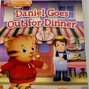 Peut inclure: Couverture de livre pour enfants intitul&eacute;e "Daniel Goes Out for Dinner" de la s&eacute;rie "Daniel Tiger's Neighborhood". La couverture pr&eacute;sente Daniel Tiger dans un sweat &agrave; capuche rouge et un serveur tenant un plateau de nourriture. L'arri&egrave;re-plan montre un restaurant.