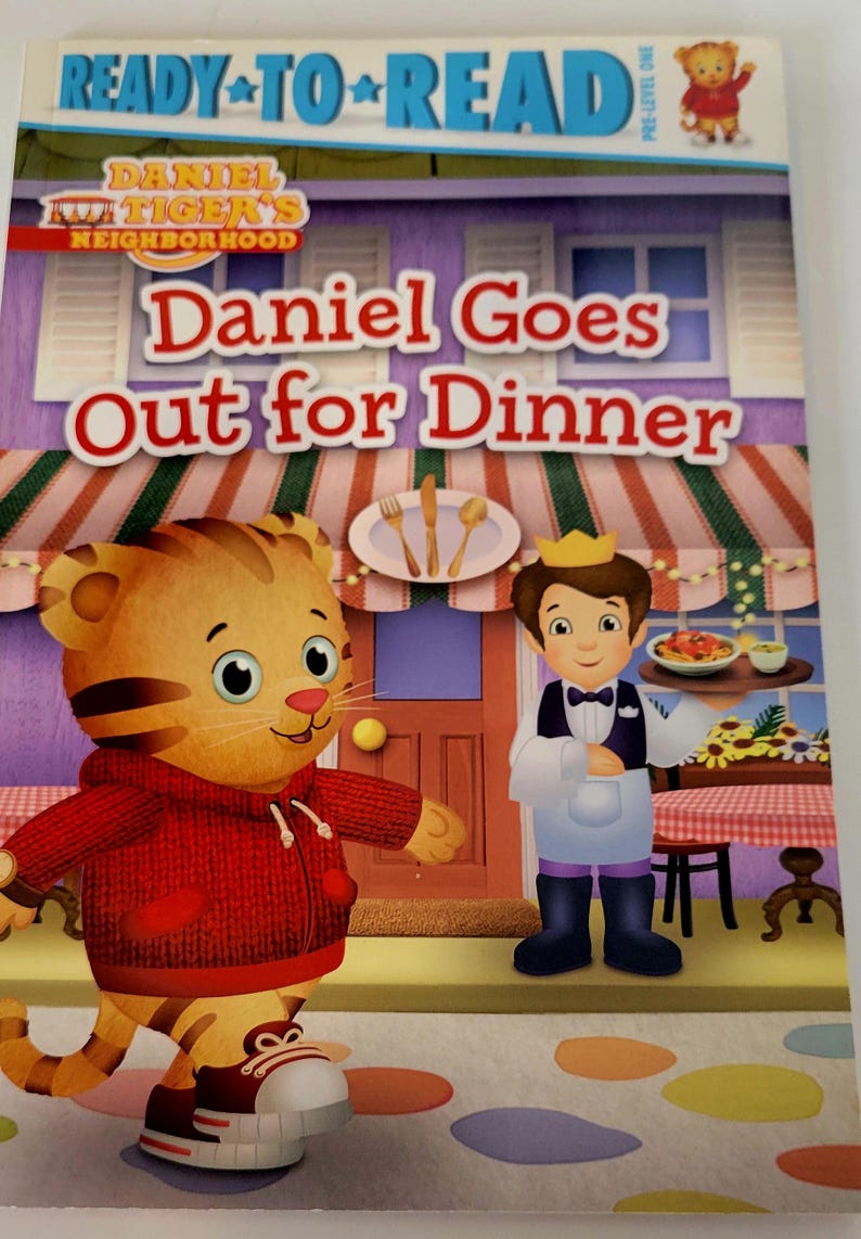 Peut inclure: Couverture de livre pour enfants intitul&eacute;e "Daniel Goes Out for Dinner" de la s&eacute;rie "Daniel Tiger's Neighborhood". La couverture pr&eacute;sente Daniel Tiger dans un sweat &agrave; capuche rouge et un serveur tenant un plateau de nourriture. L'arri&egrave;re-plan montre un restaurant.