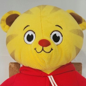 Peut inclure: Une peluche repr&eacute;sentant le visage d'un tigre jaune avec des rayures marron, de grands yeux blancs et un nez rouge. Le tigre porte un sweat &agrave; capuche rouge avec un cordon blanc. Le jouet est con&ccedil;u pour ressembler au personnage Daniel le Tigre.