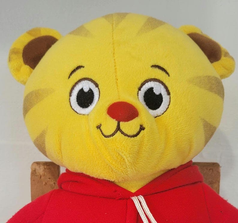 Peut inclure: Une peluche repr&eacute;sentant le visage d'un tigre jaune avec des rayures marron, de grands yeux blancs et un nez rouge. Le tigre porte un sweat &agrave; capuche rouge avec un cordon blanc. Le jouet est con&ccedil;u pour ressembler au personnage Daniel le Tigre.