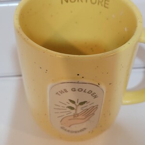 May include: Yellow ceramic mug with the word "NURTURE" printed on the rim. The mug features a graphic of a hand holding a plant with the text "THE GOLDEN GARDENER". The mug has a speckled finish.