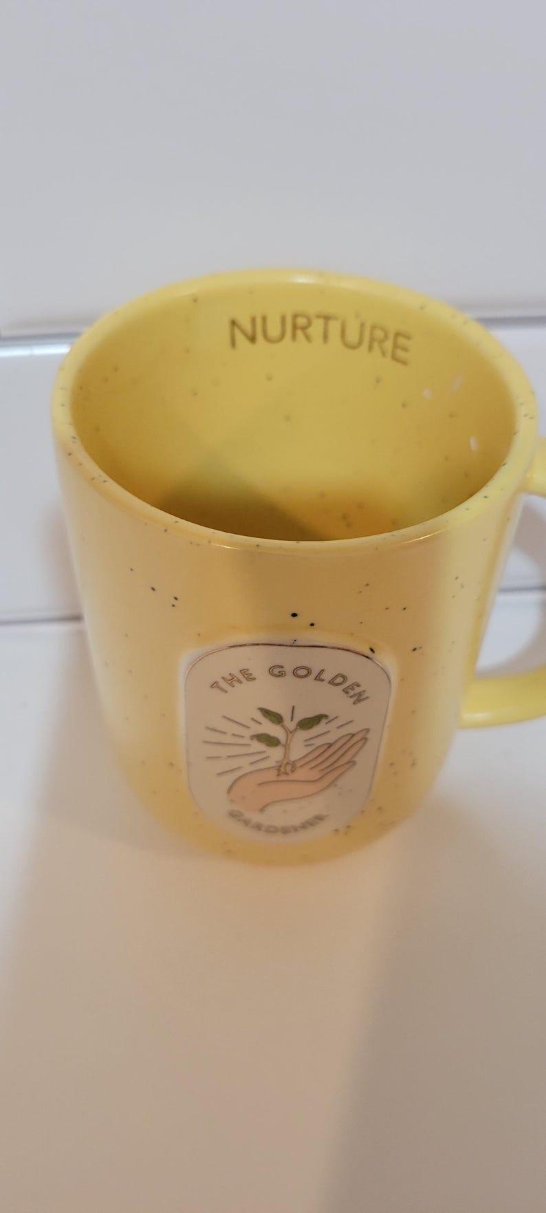 May include: Yellow ceramic mug with the word "NURTURE" printed on the rim. The mug features a graphic of a hand holding a plant with the text "THE GOLDEN GARDENER". The mug has a speckled finish.