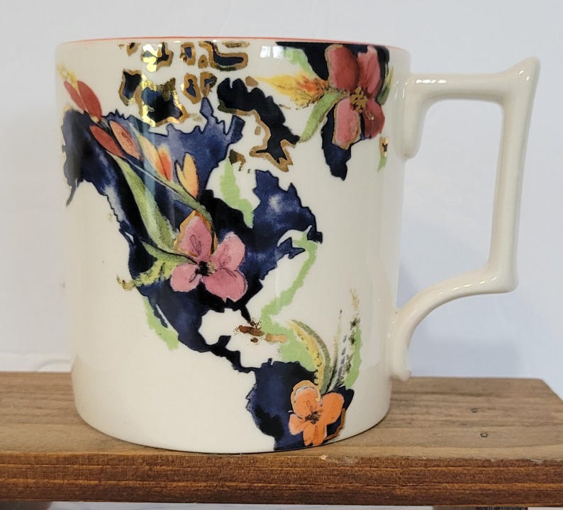 Pu&ograve; includere: Tazza in ceramica color crema con un disegno a mappa. La mappa &egrave; principalmente blu navy, con accenti dorati e illustrazioni floreali colorate. Il manico ha una forma angolare unica. Il bordo &egrave; rosa.