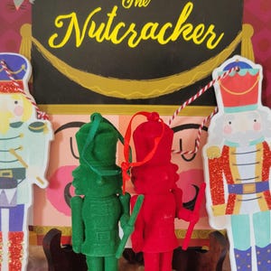 May include: The Nutcracker-themed Christmas decorations. Two plush nutcracker ornaments, one green and one red, are in front of a backdrop with the words "The Nutcracker". Additional cardboard nutcracker figures are on either side.