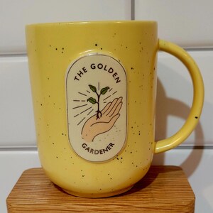 May include: Yellow ceramic mug with black speckles and a curved handle. The mug features an oval design with the words "THE GOLDEN GARDENER" and an illustration of a hand holding a sprouting plant.