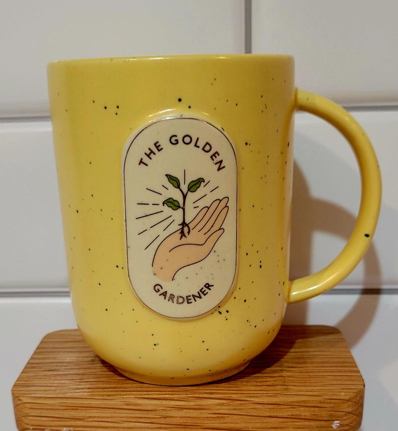 May include: Yellow ceramic mug with black speckles and a curved handle. The mug features an oval design with the words "THE GOLDEN GARDENER" and an illustration of a hand holding a sprouting plant.