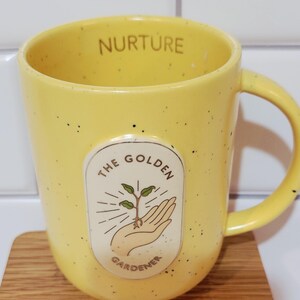 May include: Yellow ceramic mug with black speckles and a handle. The mug features the text "NURTURE" on the rim and an oval graphic with a hand holding a sprouting plant, with the text "THE GOLDEN GARDENER".