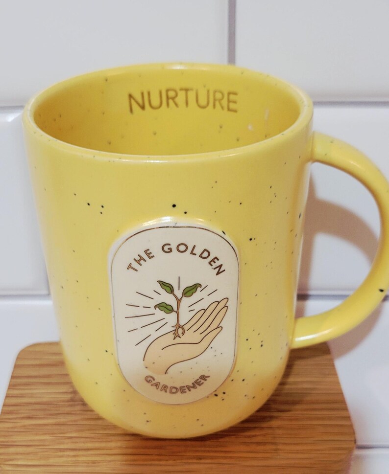 May include: Yellow ceramic mug with black speckles and a handle. The mug features the text "NURTURE" on the rim and an oval graphic with a hand holding a sprouting plant, with the text "THE GOLDEN GARDENER".