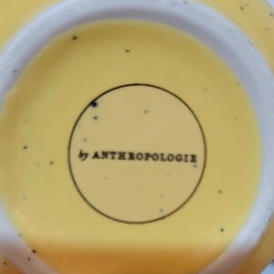 May include: Close-up of the bottom of a yellow ceramic bowl with a white rim. The bowl has small black speckles and the text "by ANTHROPOLOGIE" is printed in a black circle.