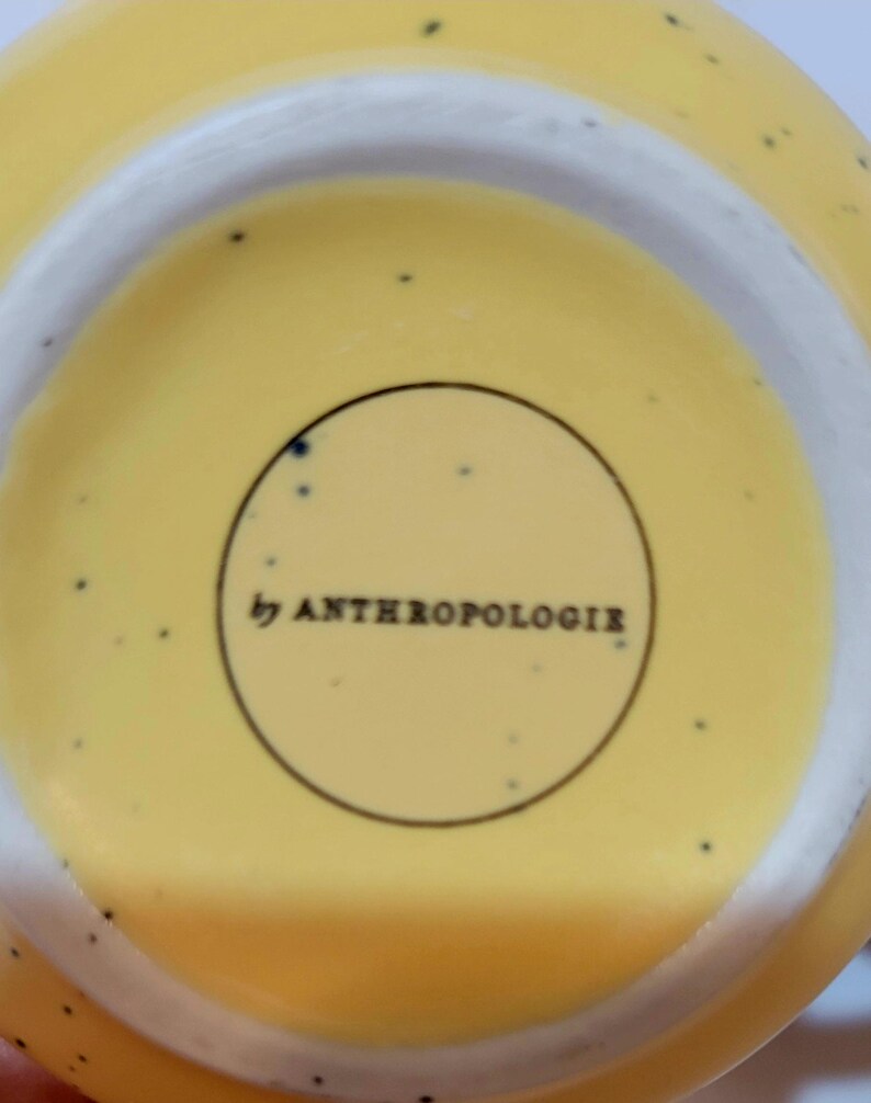 May include: Close-up of the bottom of a yellow ceramic bowl with a white rim. The bowl has small black speckles and the text "by ANTHROPOLOGIE" is printed in a black circle.