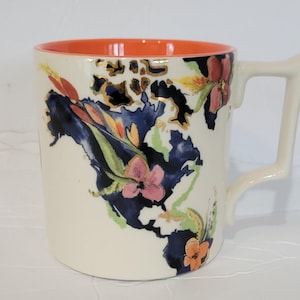 Pu&ograve; includere: Tazza in ceramica color crema con interno arancione brillante. Presenta un design floreale con fiori rosa, arancioni e blu, impreziosito da dettagli dorati. Il manico ha una forma angolare unica.