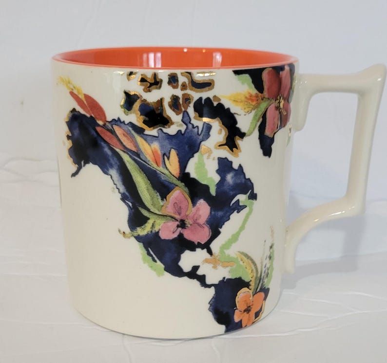 Pu&ograve; includere: Tazza in ceramica color crema con interno arancione brillante. Presenta un design floreale con fiori rosa, arancioni e blu, impreziosito da dettagli dorati. Il manico ha una forma angolare unica.
